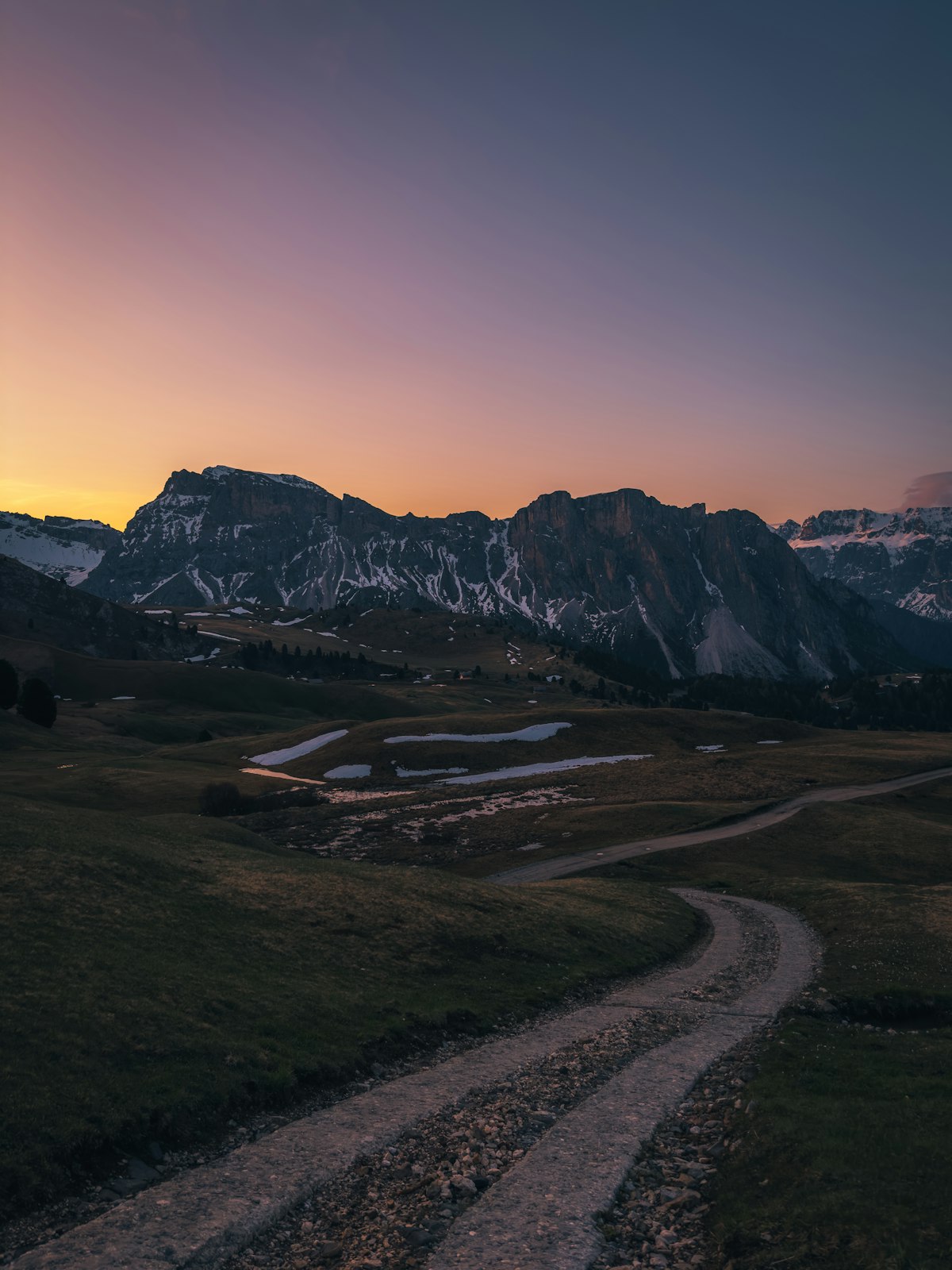 Un sentiero sterrato attraversa un altopiano erboso verso le creste rocciose al tramonto